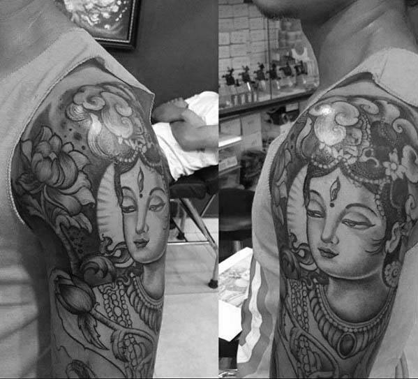 Black and grey Krishna tattoo on the upper arm featuring a serene facial expression, floral elements, and ornamental jewelry with cloud details above.