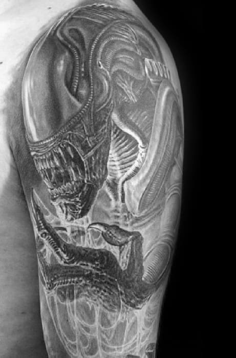 Highly detailed black and grey Xenomorph tattoo on upper arm, showcasing the alien's sharp teeth, elongated head, and clawed hand.