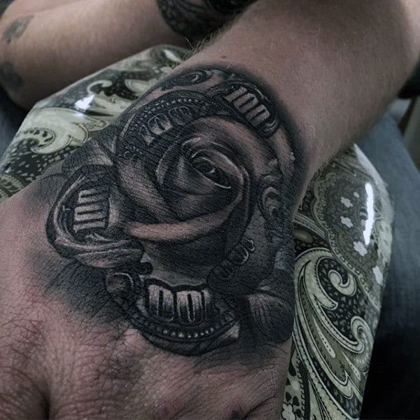 A black and gray shaded rose tattoo on the hand, with intricate details, combined with elements of money, giving a striking and bold appearance
