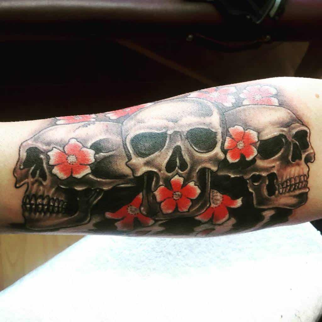 Tattoo of three detailed skulls with red flowers, symbolizing 'Hear No Evil, See No Evil, Speak No Evil,' in a bold black and gray style with vibrant accents