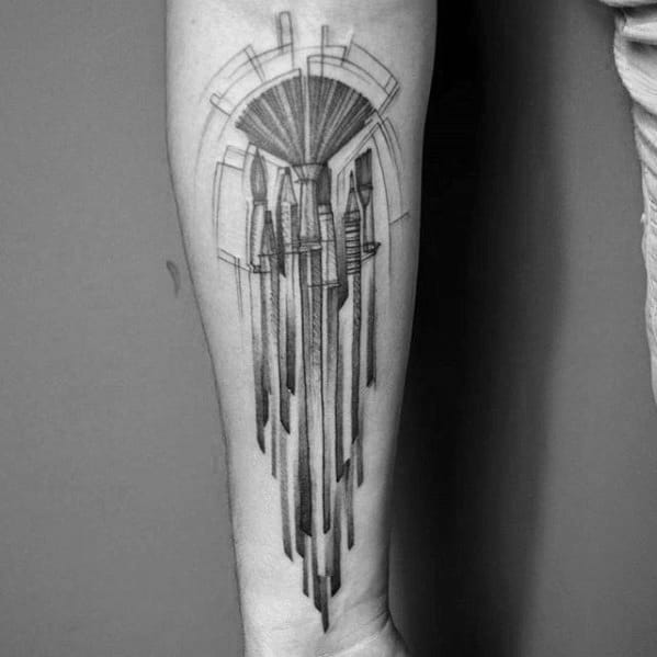 A geometric tattoo of art tools, showcasing intricate brushes and a distinctive pencil, elegantly adorns a person's forearm