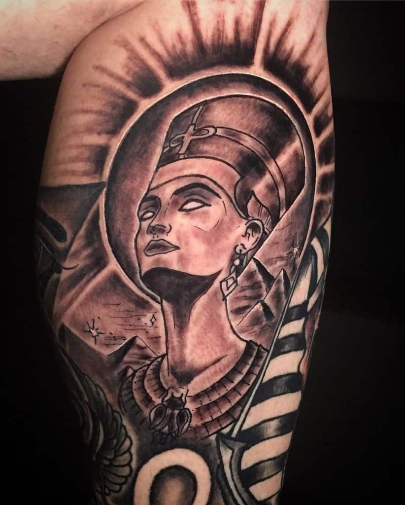 Tattoo of Nefertiti with a headdress, necklace, and ankh symbol against a dark background