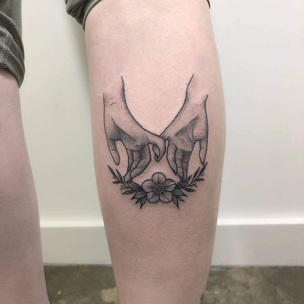 Shaded pinky promise tattoo with two detailed hands linked above a floral design, placed on the back of the calf