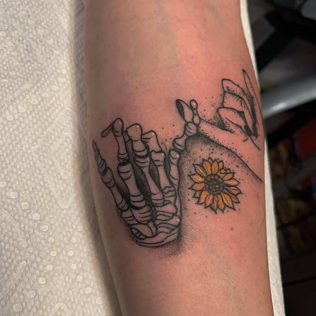 Tattoo of a skeleton hand and human hand making a pinky promise, with a sunflower nearby, on a forearm