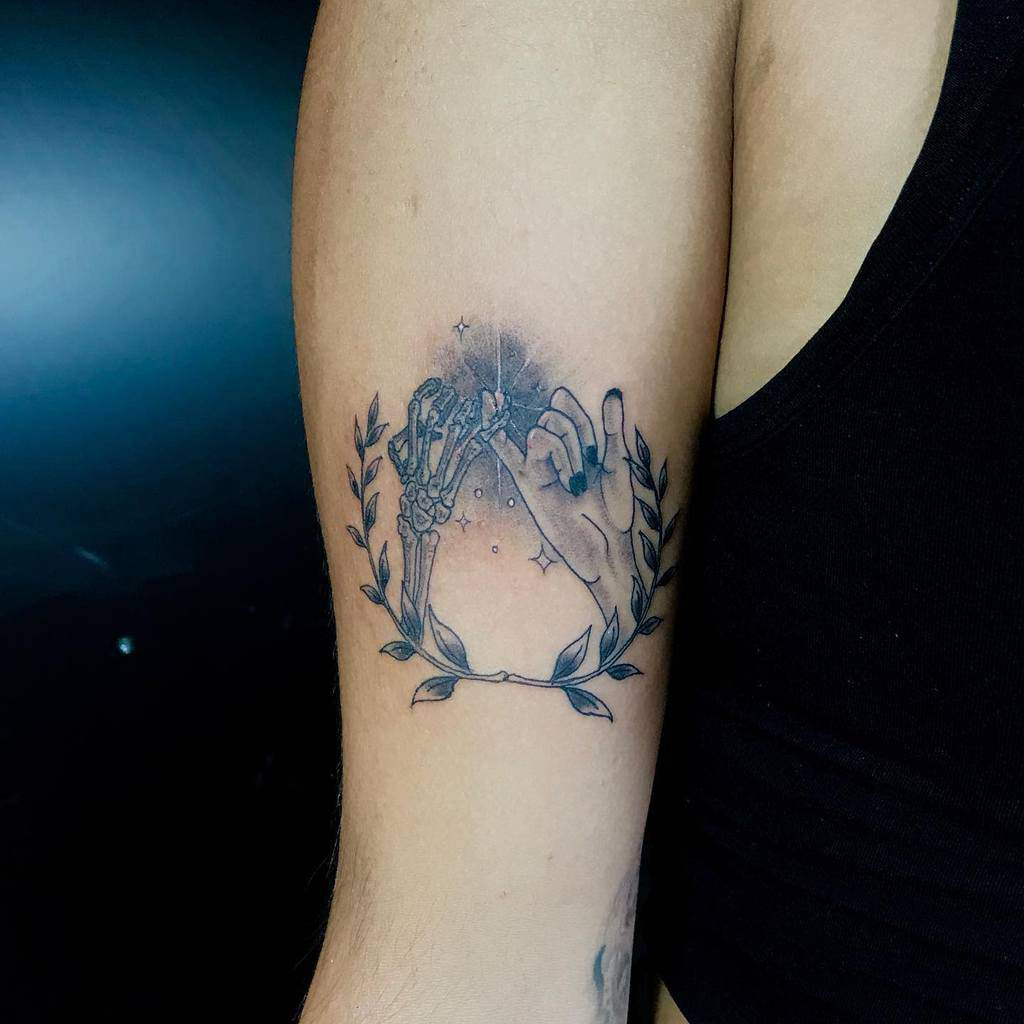 Tattoo of intertwined skeletal and human hands making a pinky promise, surrounded by laurel branches on an upper arm