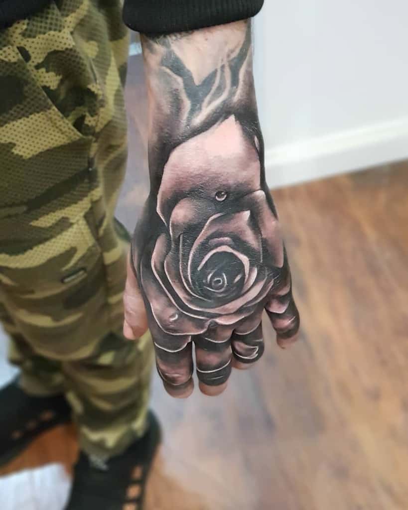 A detailed black and gray shaded rose tattoo covering the hand, featuring realistic texture and highlights on the petals