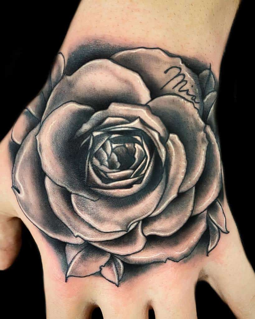A striking black and gray tattoo of a blooming rose adorns the back of a hand, showcasing its intricate details