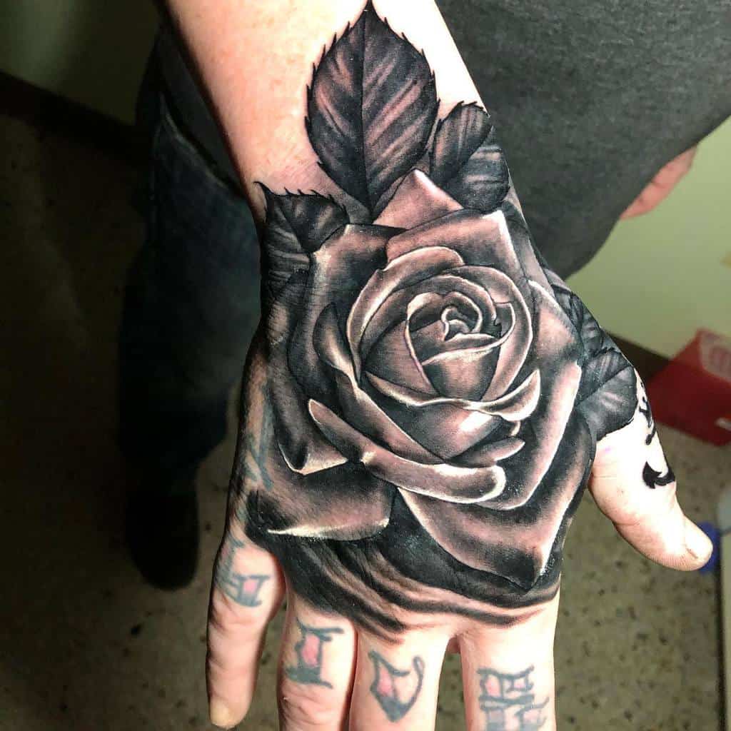A black and white rose tattoo gracefully adorns the back of a hand, its intricate petals flowing seamlessly into elegantly tattooed letters on the fingers