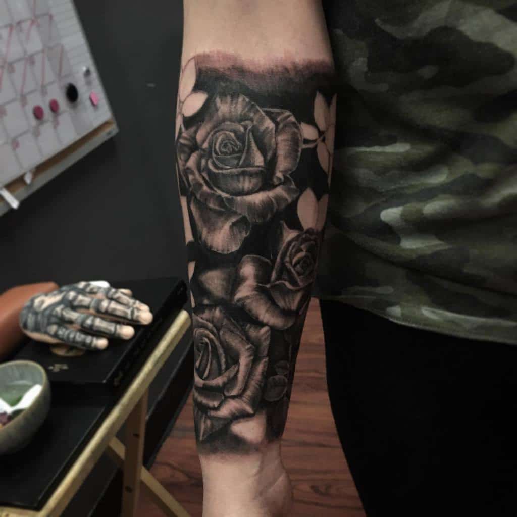 A realistic black and gray shaded tattoo featuring large roses and detailed leaves, covering the forearm with soft shading for depth and contrast