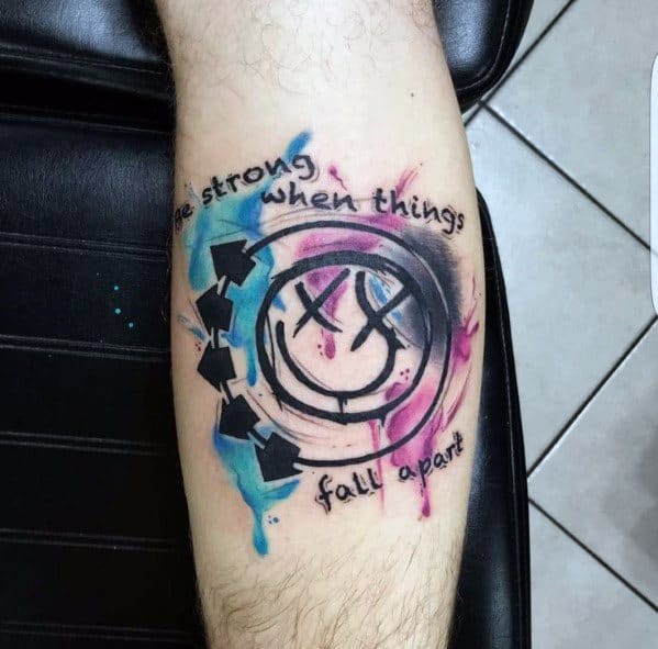 Tattoo of the Blink-182 smiley face logo with blue and pink watercolor splashes, accompanied by the phrase "Be strong when things fall apart" on the forearm.