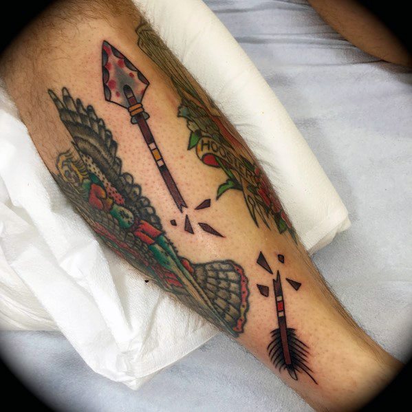 Neo-traditional broken arrow tattoo on the leg with red and black accents, surrounded by flying fragments and bold illustrative elements.