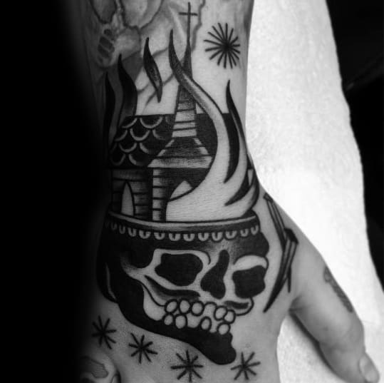 Bold blackwork tattoo on the hand featuring a burning church rising from a skull. Surrounded by stars and dotwork elements for added texture.