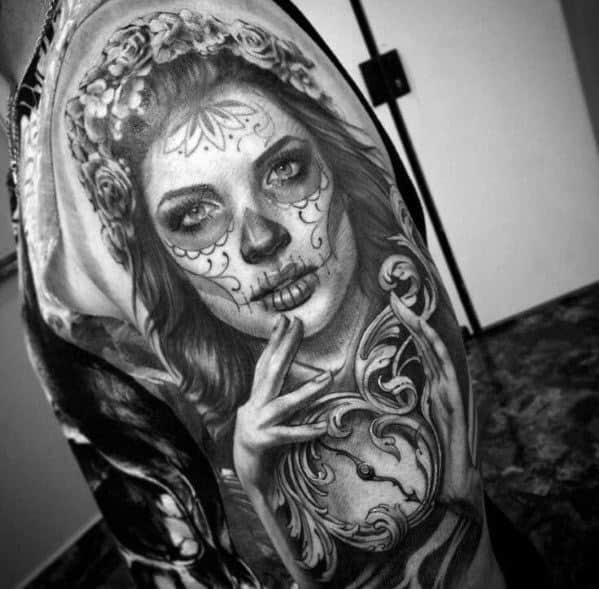 La Catrina tattoo of a woman with sugar skull makeup, floral crown, and ornate frame design on the shoulder in black and grey style.
