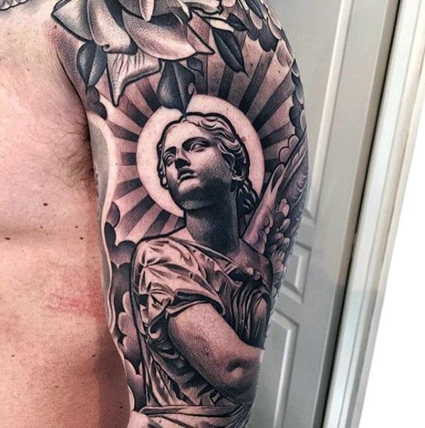 A black and grey tattoo on the upper arm featuring an angel with large wings, set against a radiant sunburst and surrounded by detailed flowers.