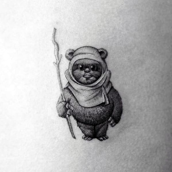 Tiny black and grey Ewok tattoo with soft shading, holding a crooked staff and wearing a simple hood with a calm, curious expression.