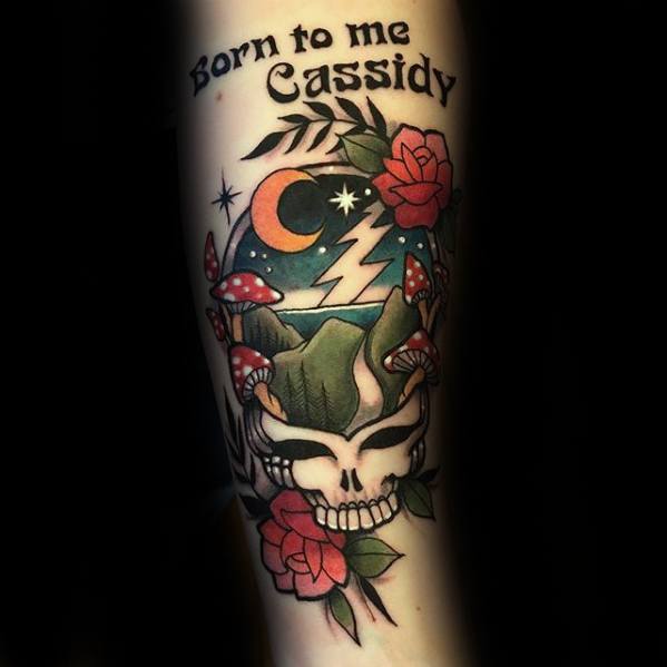 Grateful Dead inspired skull tattoo with mountains, lightning, moonlit sky, roses, mushrooms, and the text “Born to me Cassidy” above.
