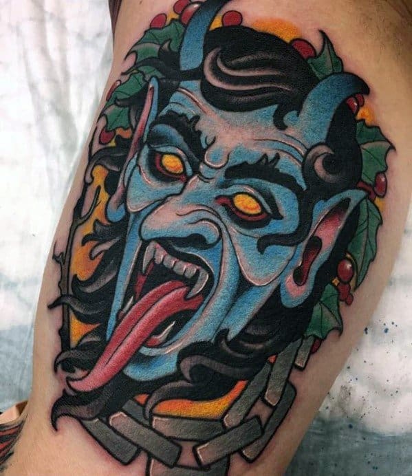Bold Krampus tattoo featuring a blue demon face with glowing orange eyes, long red tongue, chains, and holly leaves for a festive touch.