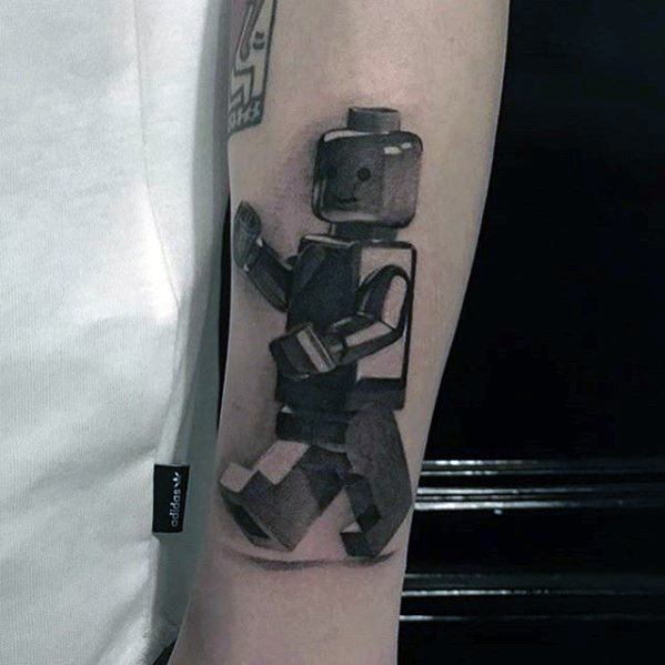 Realistic black and grey tattoo of a walking Lego minifigure, featuring strong shadows and a 3D effect on the inner forearm.