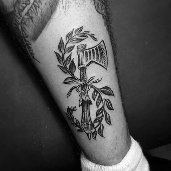 A pencil-drawn tattoo of an axe elegantly wrapped in olive branches adorns a person's lower leg