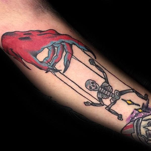 Colorful puppet tattoo of a red, claw-like hand controlling a dancing skeleton with strings, blending dark humor with a bold graphic style.