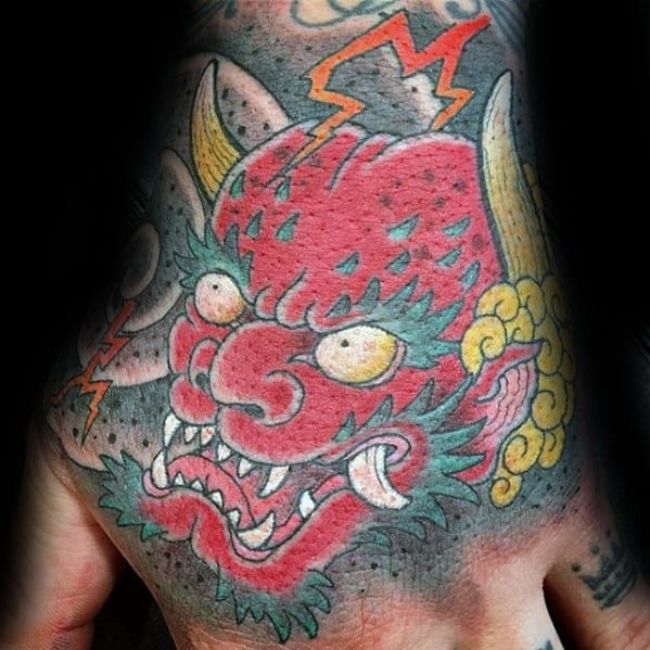 Fierce Raijin hand tattoo with red thunder god face, yellow horns, lightning bolts, and bold Japanese-style detailing.