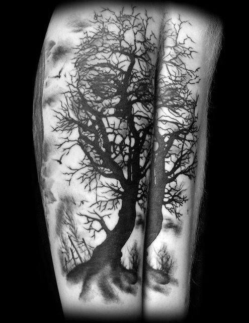 Split-leg tattoo of intertwining trees with branches forming a hidden skull, set in a foggy forest scene with flying birds.
