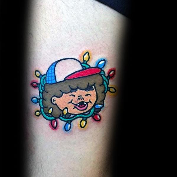 Cute cartoon-style tattoo of Dustin from Stranger Things, smiling and framed by colorful Christmas lights.