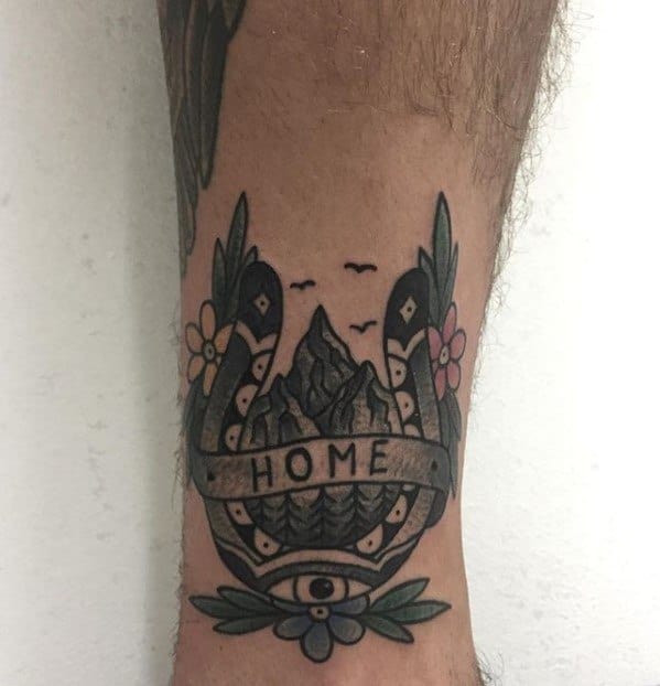 Traditional style tattoo of a mountain scene framed by a horseshoe, flowers, and leaves, with a banner reading “HOME” and an all-seeing eye.