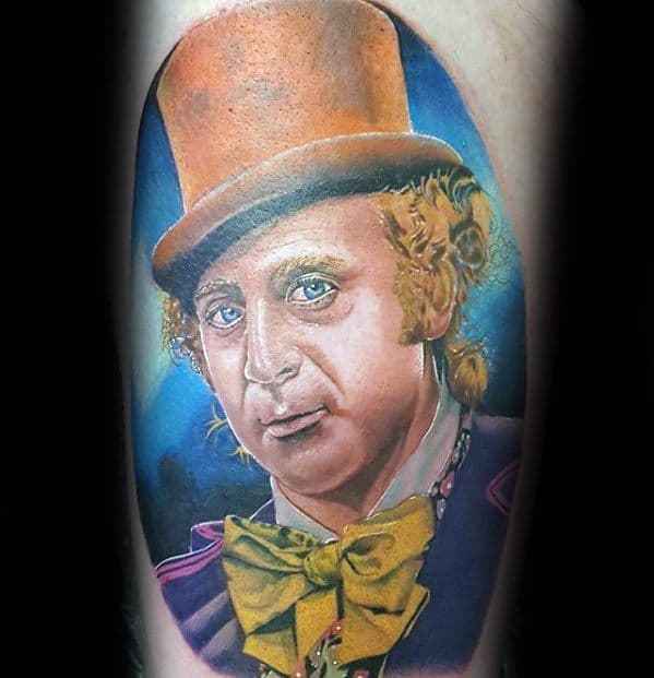 Vivid Willy Wonka tattoo with a warm orange hat, curly blond hair, yellow bow tie, and a rich blue background highlighting the portrait.