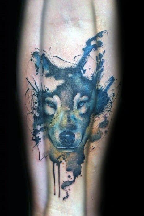 Watercolor wolf tattoo with piercing eyes, abstract ink splatters, and sketch-style lines in blue, yellow, and black on the forearm.