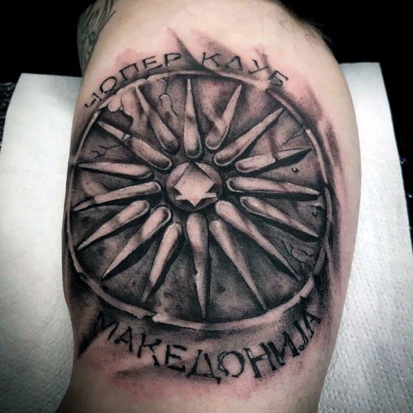 Tattoo of the Vergina Sun symbol with bold shading, surrounded by Cyrillic text reading “ЧОПЕР КЛУБ МАКЕДОНИЈА” on the upper arm.