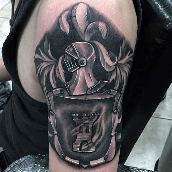 Black and grey upper arm tattoo of a medieval knight helmet above a shield featuring a castle tower emblem, framed by ornate flourishes.