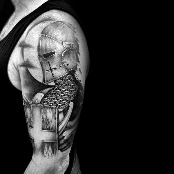 Realistic black and grey upper arm tattoo of a medieval knight in chainmail and helmet, holding a shield with a bold cross symbol.