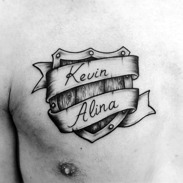 Chest tattoo of a wooden shield wrapped in a ribbon banner, featuring the names “Kevin” and “Alina” in elegant script.
