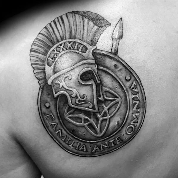 Black and grey shoulder tattoo of a Spartan helmet over a circular shield with a pentagram, inscribed with “FAMILIA ANTE OMNIA” and Roman numerals.