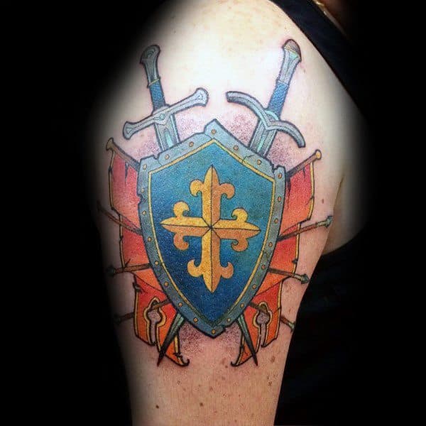 Colorful upper arm tattoo of a blue and gold medieval shield with a fleur-de-lis design, crossed swords, and red banners in the background.