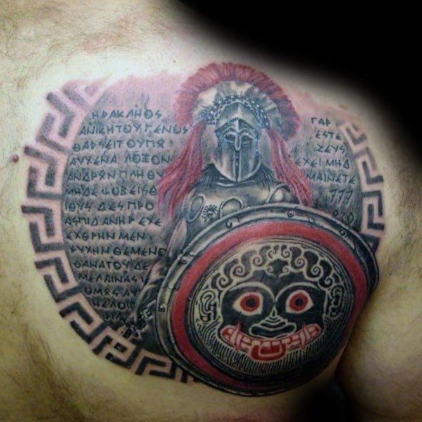 Bold chest tattoo of an ancient Greek warrior with red-plumed helmet and ornate shield featuring a Gorgon face, surrounded by Greek text and a meander border.