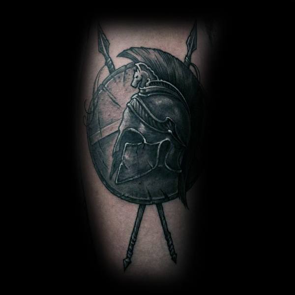 Dark-toned tattoo of a Spartan helmet and shield with crossed spears behind, creating a powerful symbol of warrior spirit and ancient strength.