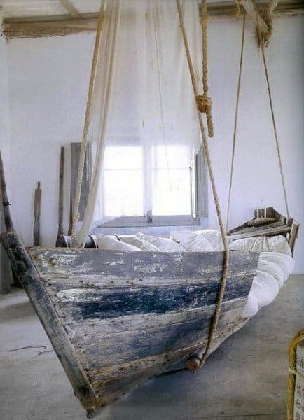 Rustic boat-shaped hanging bed with rope supports and white bedding in a nautical-themed room.