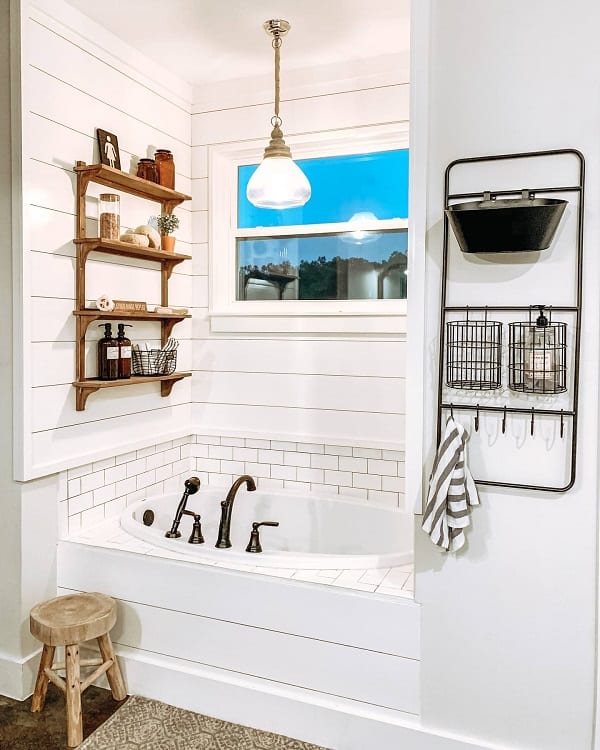 Farmhouse bathroom with white shiplap walls, built-in bathtub, wooden shelves, window, and pendant light
