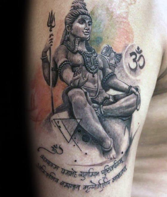 Upper arm tattoo of Lord Shiva seated with a trident, accented by geometric elements, Om symbols, Sanskrit script, and soft watercolor shading.
