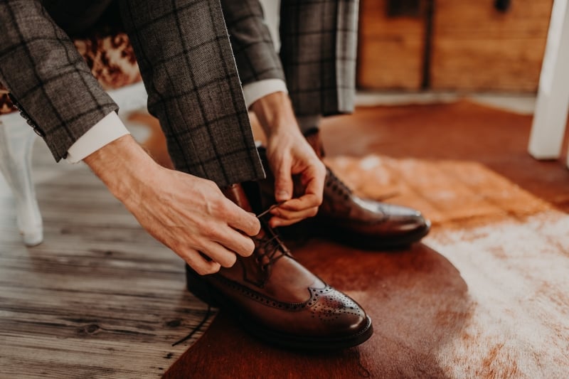 A person in a grey plaid suit sits on a chair and ties the laces of their brown leather dress shoes