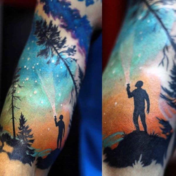 Tattoo of a person with flashlight under a colorful night sky with trees around