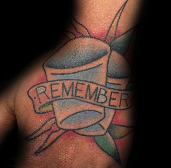 Bold shot glass tattoo with two glasses, a "REMEMBER" banner, and leaf accents, featuring a colorful and symbolic design on the hand.