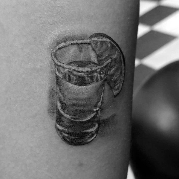 Realistic black and grey shot glass tattoo featuring a salted rim and a lime wedge, creating a classic tequila-inspired design with fine shading.