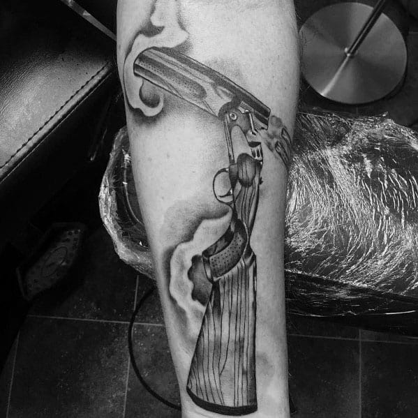 Realistic black and grey tattoo of a smoking double-barrel shotgun, featuring intricate shading, wood grain texture, and dynamic smoke effects.