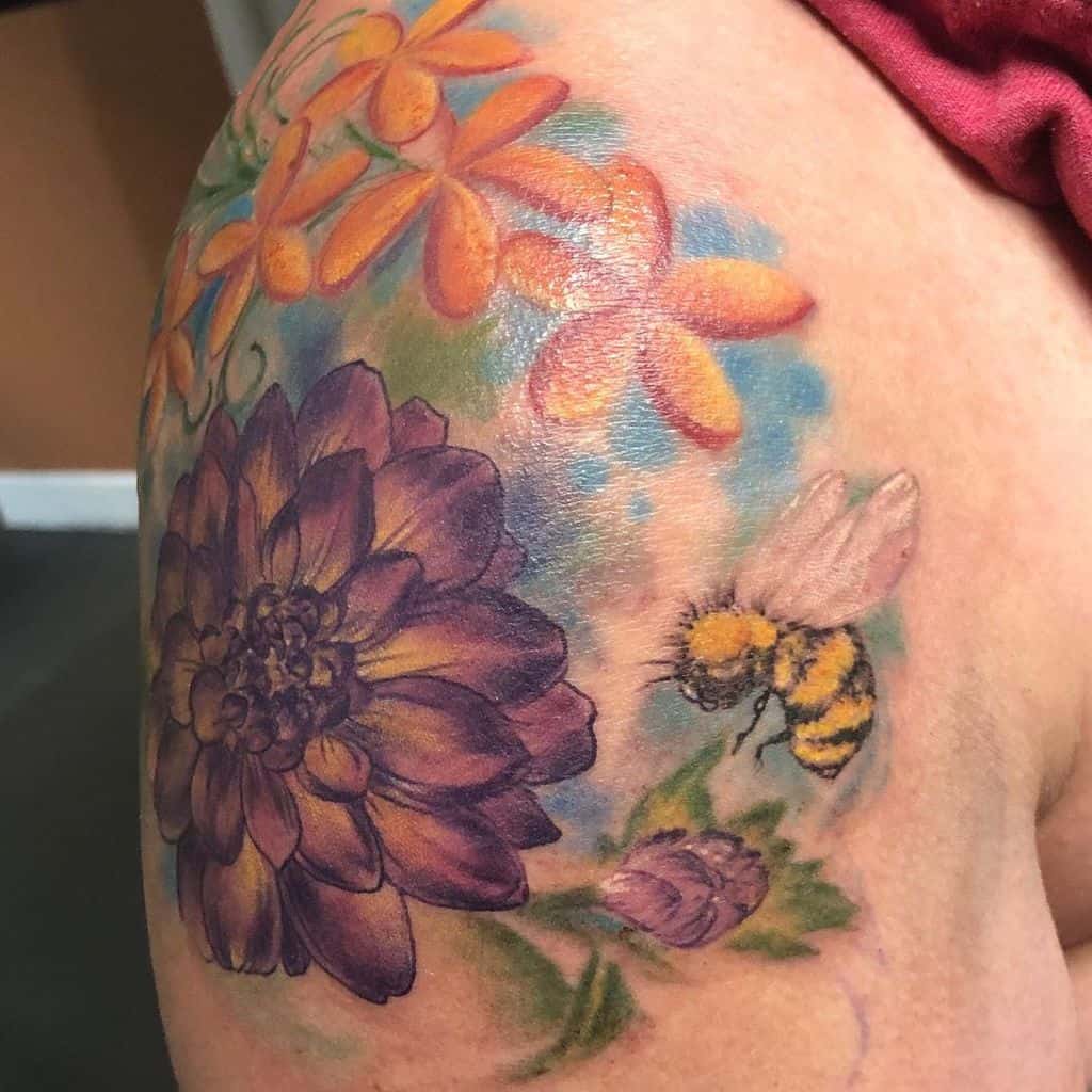 Colorful shoulder tattoo of a bee hovering near purple and orange flowers with a vibrant watercolor background