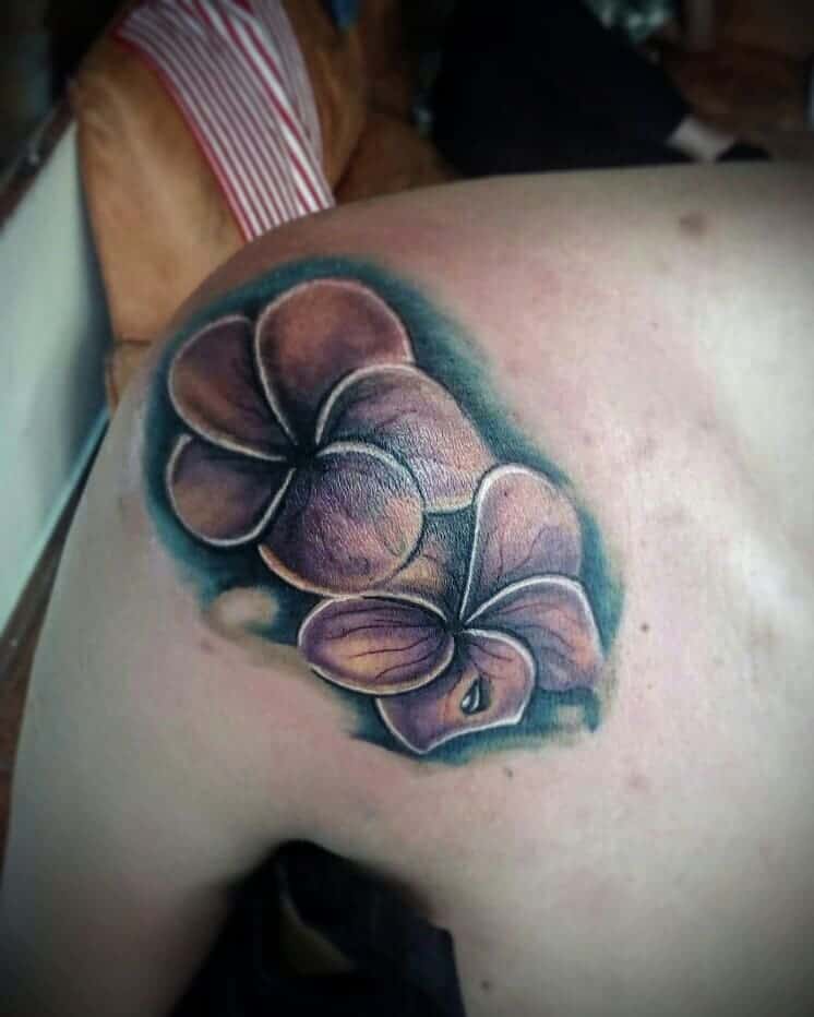 Detailed shoulder tattoo of purple plumeria flowers with soft shading and a teal background for added depth