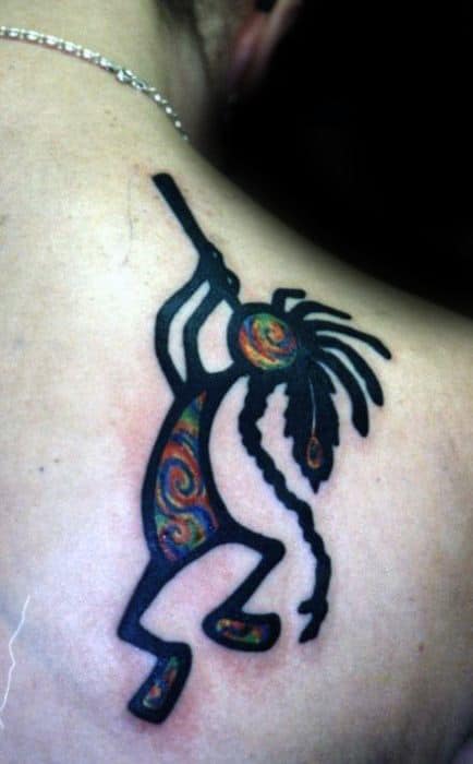 Kokopelli tattoo with swirling colors in the body and hair, contrasted by bold black outlines, creating a striking, artistic design on the shoulder.
