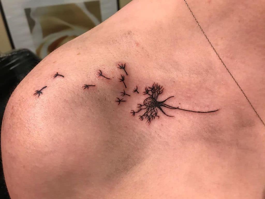 This tattoo illustrates a dandelion with seeds blowing away, symbolizing freedom and letting go. The design is simple yet powerful, placed across the shoulder, creating a flow of movement with the scattered seeds.